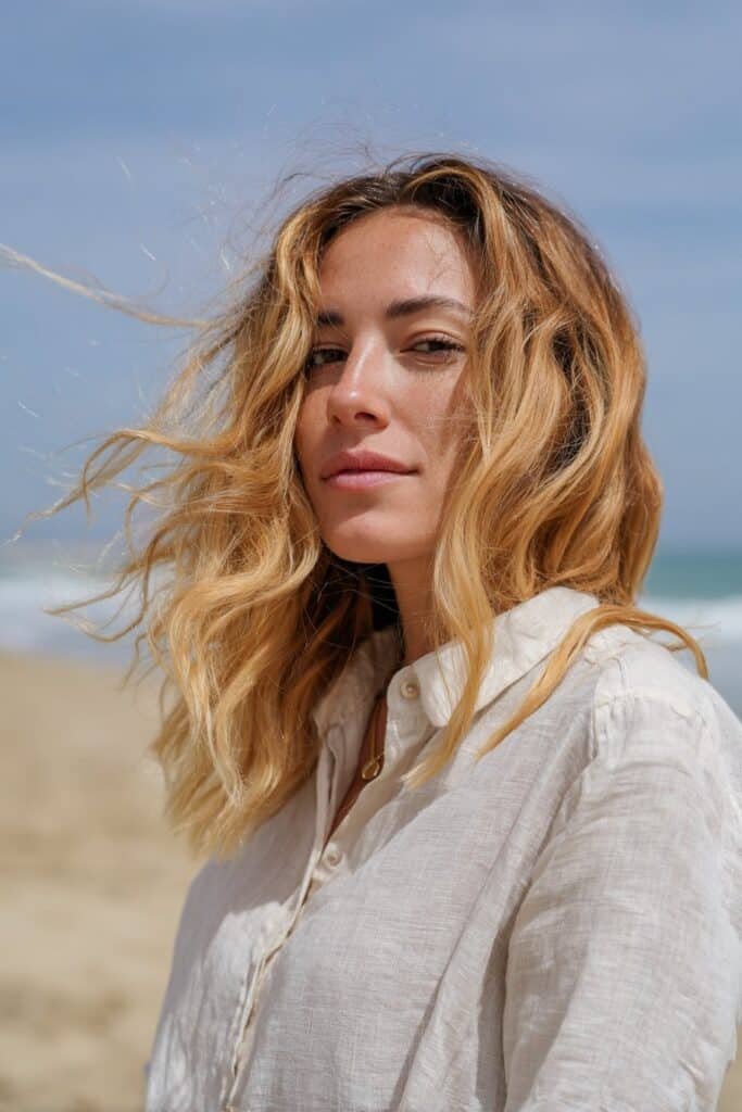 Young woman with beachy golden blonde highlights and soft waves, captured near a sandy beach with natural sunlight.