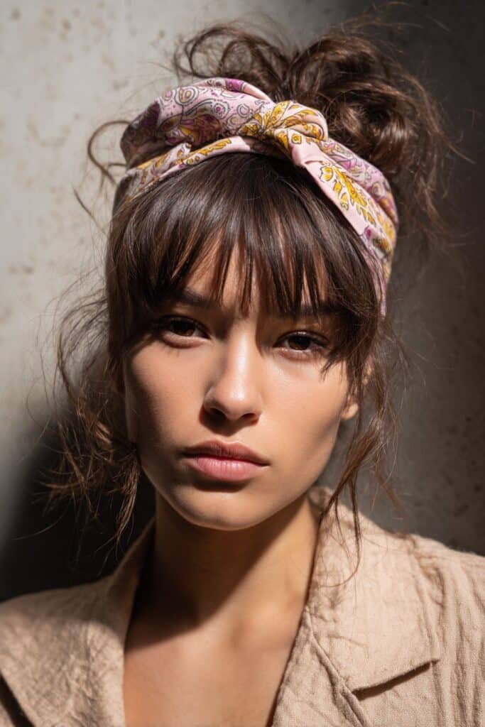 A close-up photo of a young elegant American woman with wispy bangs styled with a patterned boho bandana headband, featuring realistic hair texture under soft natural lighting, subtle makeup, warm skin tone, confident expression, fully clothed in a chic outfit, in an Instagram-style beauty shot – headband with bangs series.