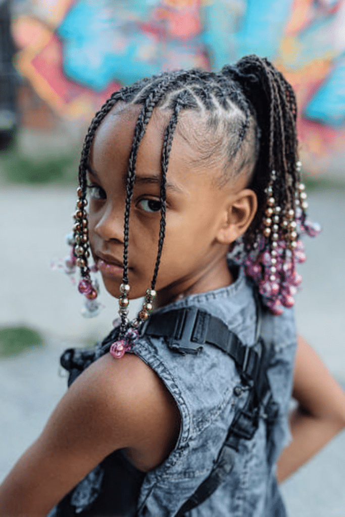 Toddler girl styled with a braided mohawk ponytail decorated with beads in an urban park.