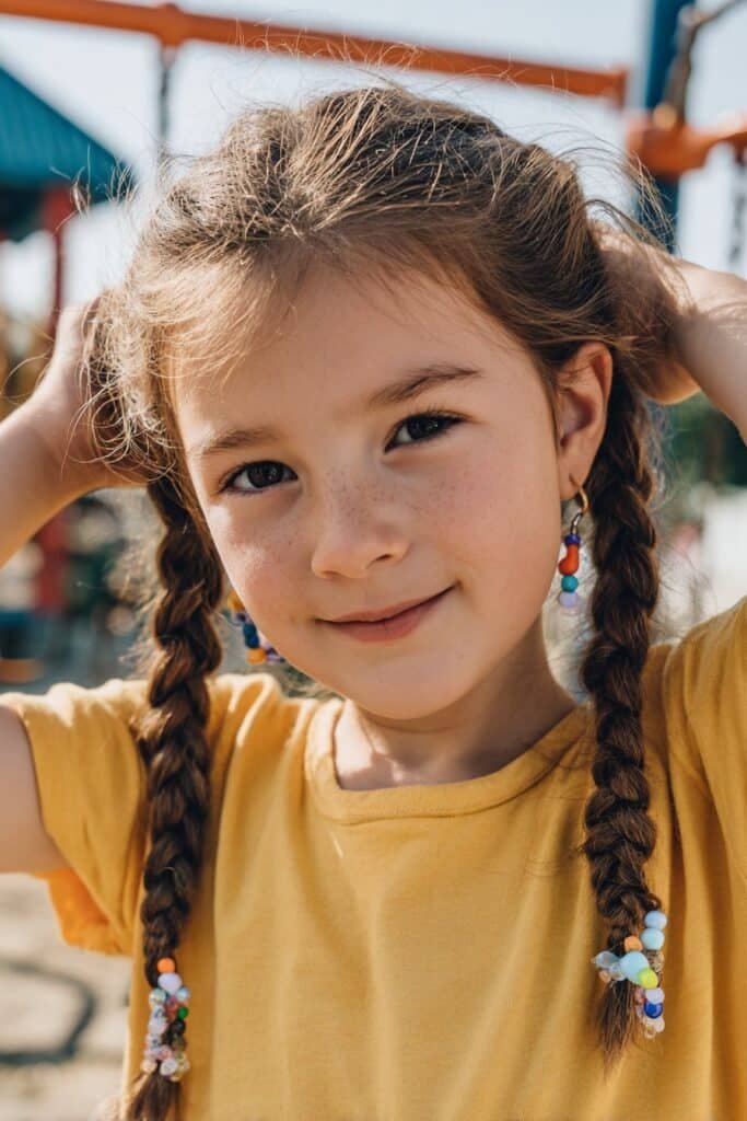 Braided pigtails with beads on an 8-year-old girl, outdoor park background, green trees, bright natural sunlight