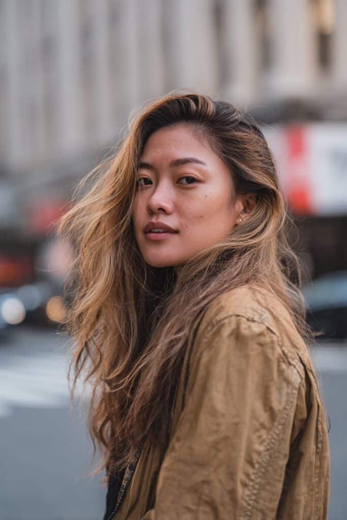 Woman in her 20s walking forward with hair flowing to showcase buttery blonde balayage highlights on a city street at golden hour.