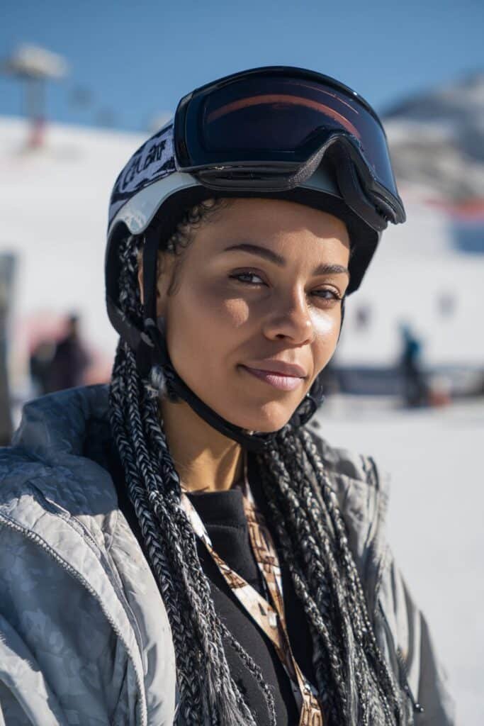 Front-facing portrait of a woman in her 20s wearing a ski helmet with cornrow braids, a protective ski hairstyle that stays flat under the helmet.