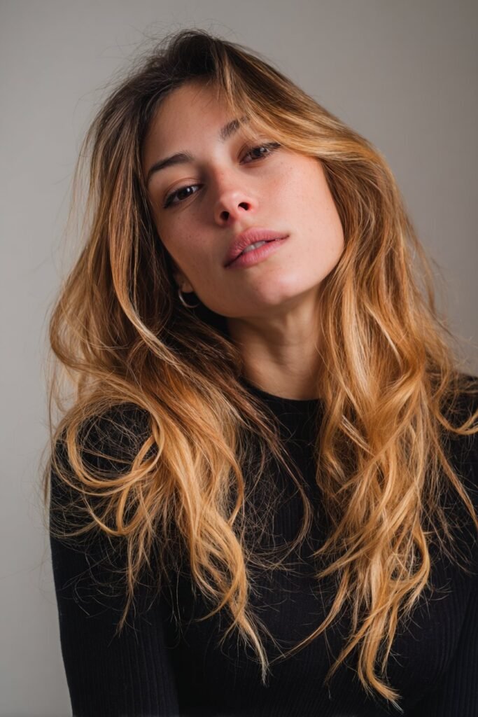 Woman in her 20s turning halfway away from the camera to display creamy blonde ombre color fading through the lengths in a fashion studio.