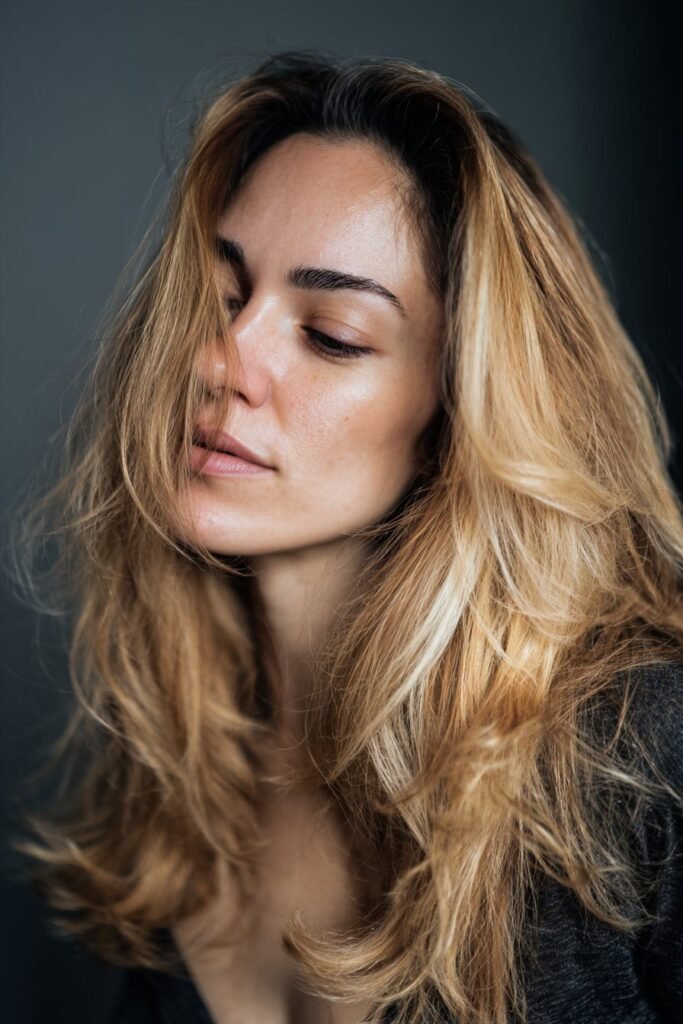 Woman in her 20s tilting her head downward to emphasize creamy blonde hair with a soft root shadow against a dark studio background.