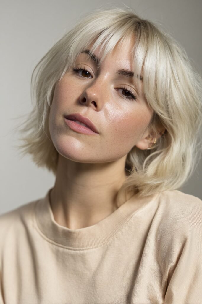 Young woman wearing a cropped platinum haircut with fringe, soft icy bangs framing the face in a clean neutral studio.