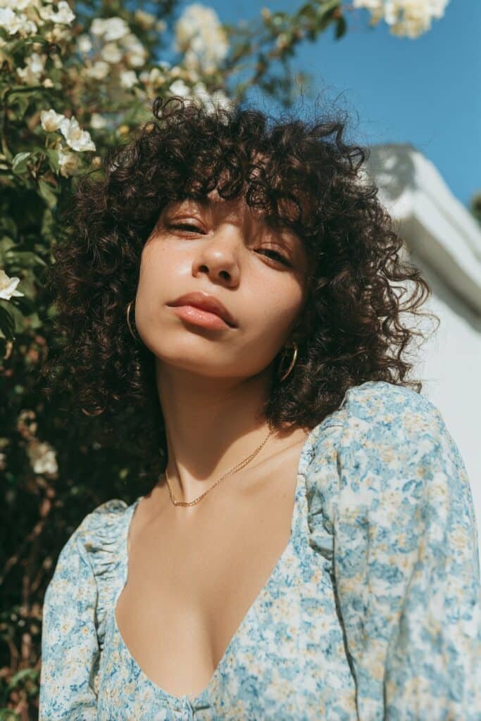 Young woman in her 20s with fine thin curly hair with curly bangs, floral dress, standing in a blooming spring garden.