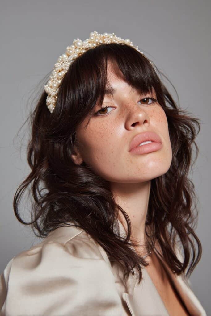 A close-up photo of a young elegant American woman with soft wavy bangs and a jeweled tiara headband in Disney princess style, featuring realistic hair texture under soft natural lighting, subtle makeup, warm skin tone, confident expression, fully clothed in a chic outfit, in an Instagram-style beauty shot – headband with bangs series.