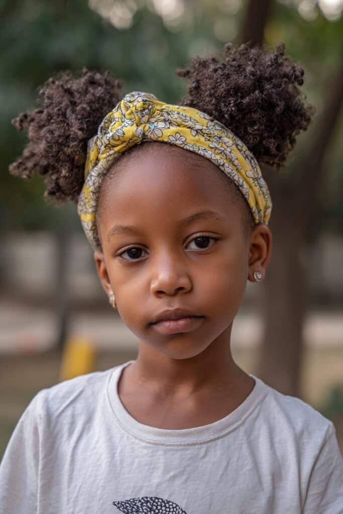 A cheerful little girl with double afro puffs wearing a soft stretchy headband, dressed in a cute outfit, smiling in a playful park with greenery in the background.