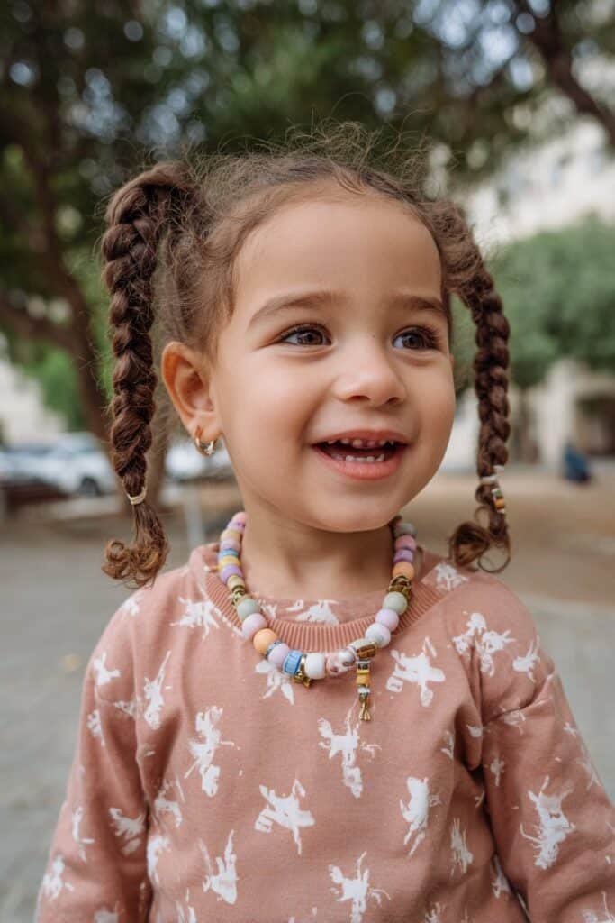 A toddler girl with detailed fishbone braids and colorful beads, photographed outdoors with a nature-inspired background.