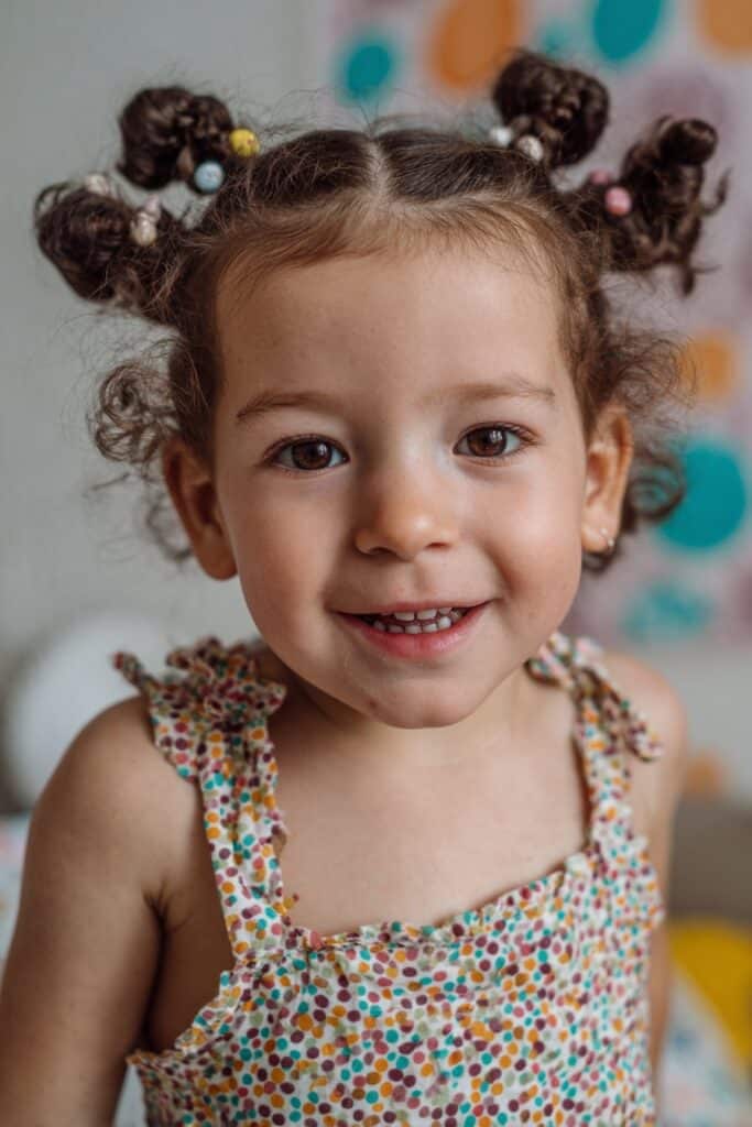 A toddler girl with four braided buns featuring matching beads, photographed in a bright party-themed setting.