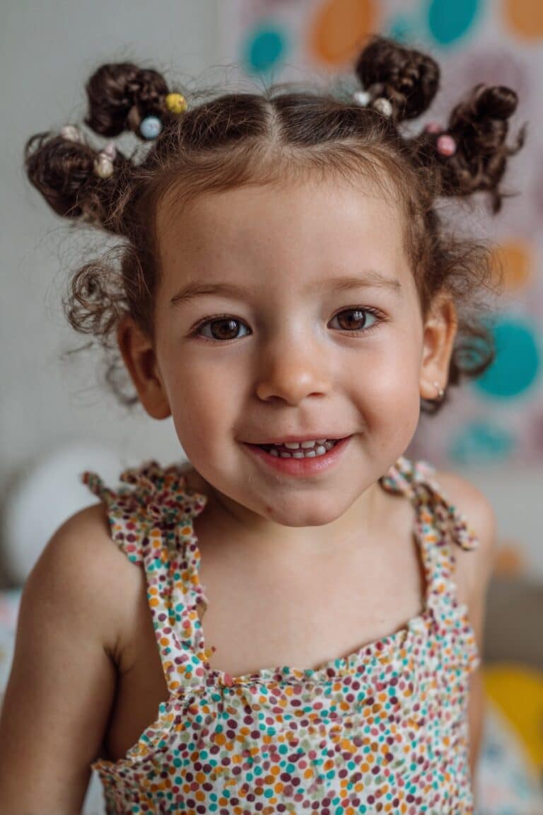 A toddler girl with four braided buns featuring matching beads, photographed in a bright party-themed setting.