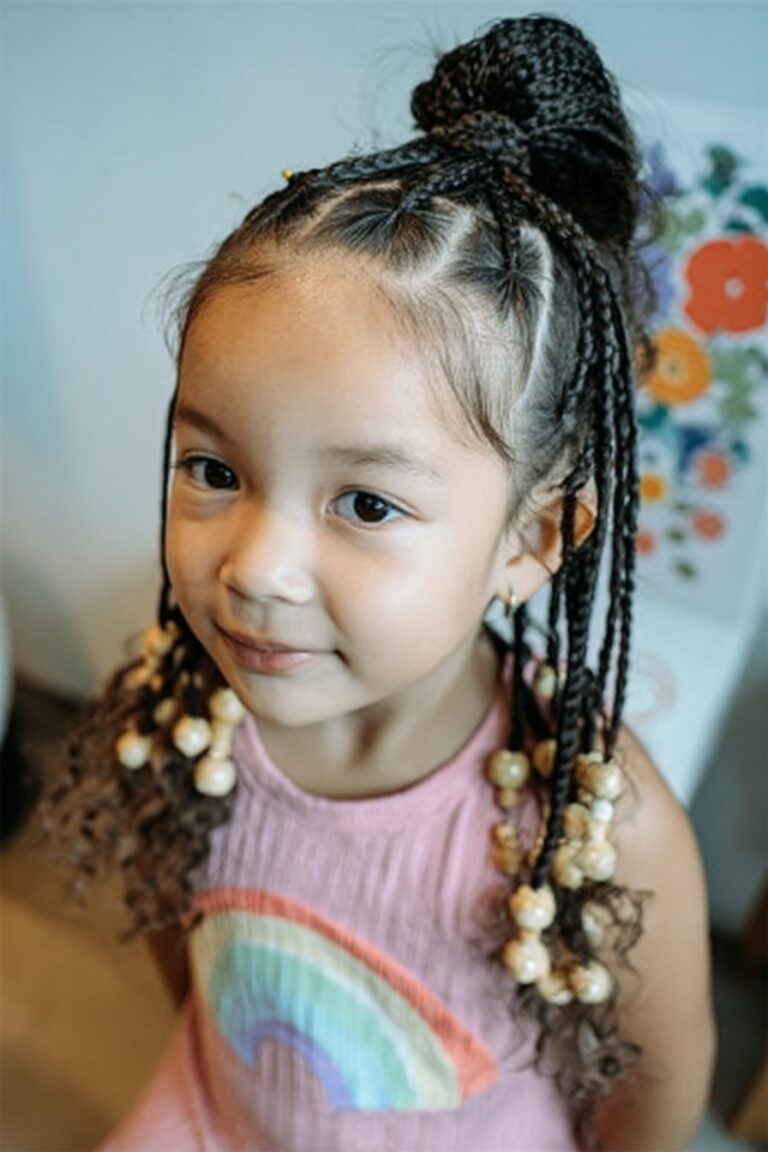 Toddler girl wearing half-up half-down braids forming a beaded ponytail in a cozy living room.
