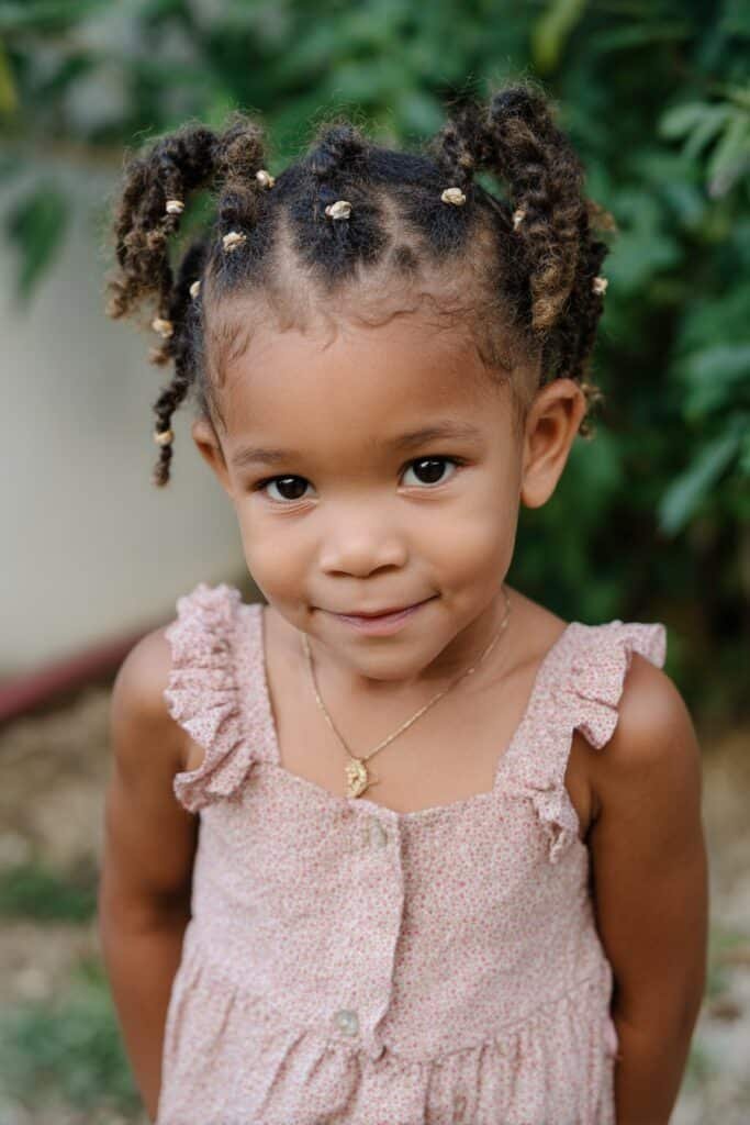 A toddler girl wearing a halo braid with small beads, photographed outdoors in a flower garden setting.