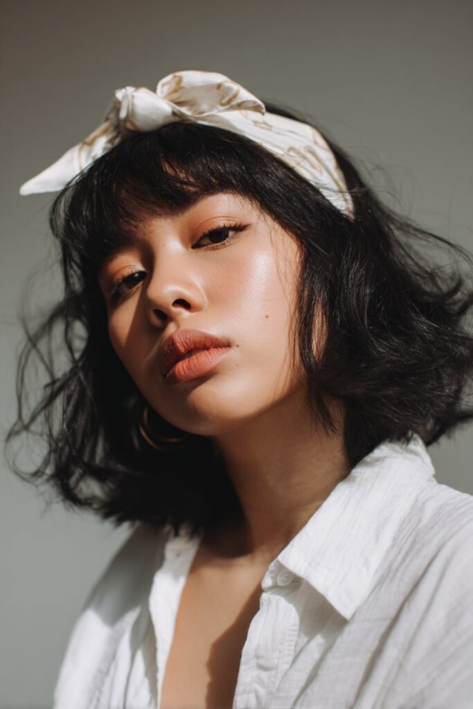 A close-up photo of a young elegant American woman with avant-garde bangs and an embellished editorial headband, featuring realistic hair texture under soft natural lighting, subtle makeup, warm skin tone, confident expression, fully clothed in a chic outfit, in an Instagram-style beauty shot – headband with bangs series.