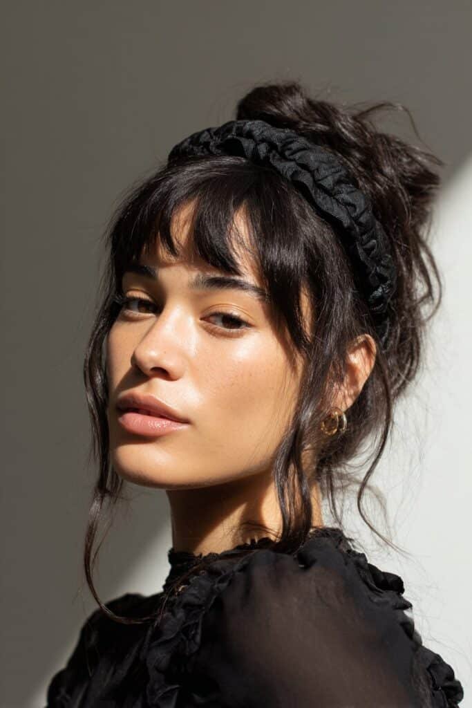 A close-up photo of a young elegant American woman with teased bangs boosted by a padded headband for volume, featuring realistic hair texture under soft natural lighting, subtle makeup, warm skin tone, confident expression, fully clothed in a chic outfit, in an Instagram-style beauty shot – headband with bangs series.