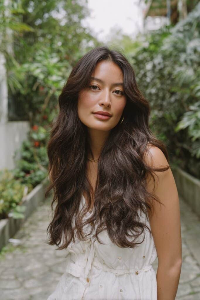 Chest-up front-facing image of a woman in her 20s with long layered hair, emphasizing flowing length and dimension in a garden pathway setting.