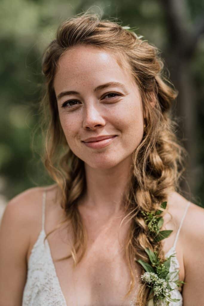 Bride in her 20s styled with a loose fishtail braid wedding hairstyle accented with greenery in a serene forest ceremony setting