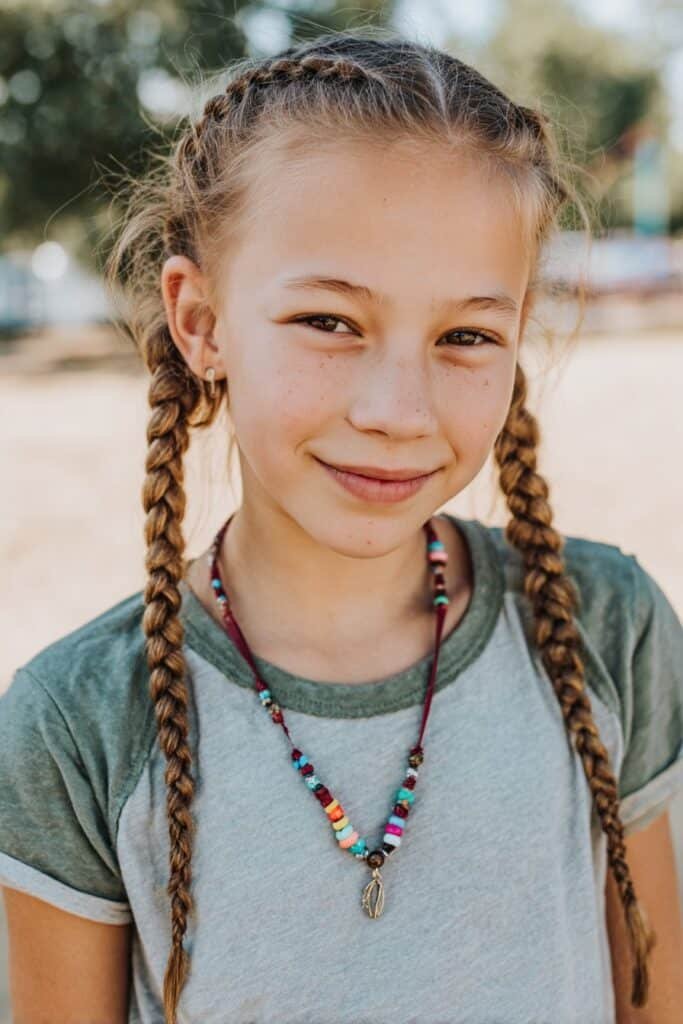 Middle part tribal braids on an 8-year-old girl with colorful beads, smiling in a bright school playground background, natural daylight, soft child-friendly look