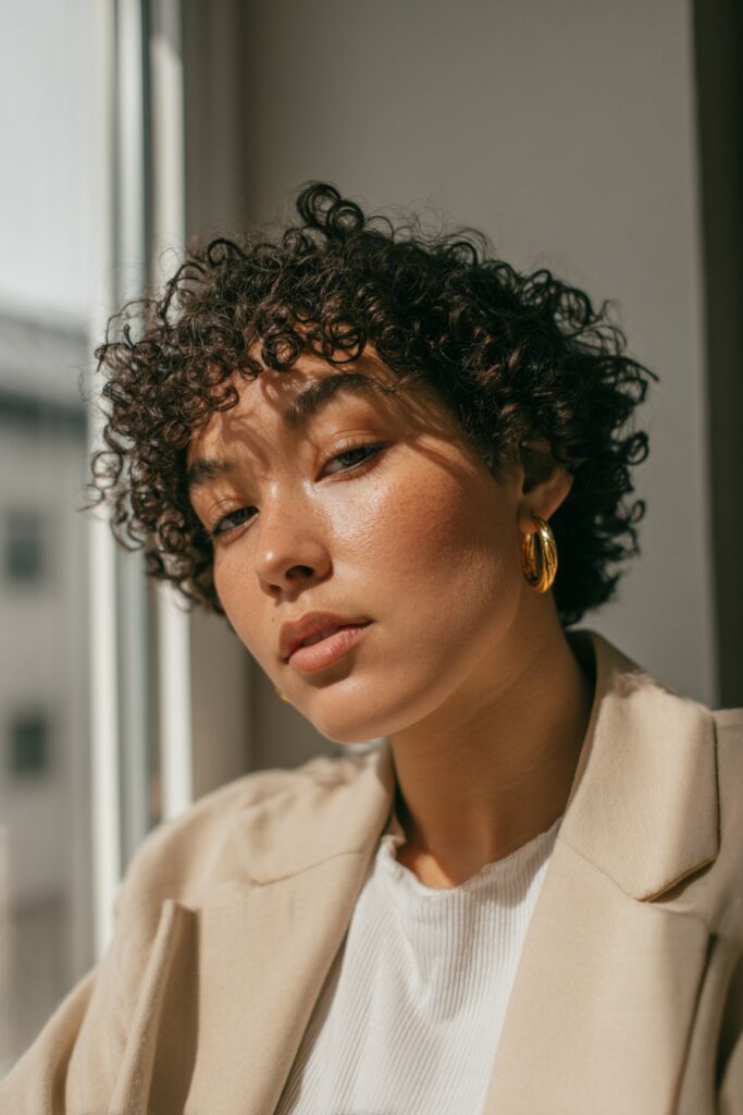 Young woman in her 20s with fine thin curly hair in a pixie cut styled with finger curls, soft makeup, light outfit, cozy indoor window setting.