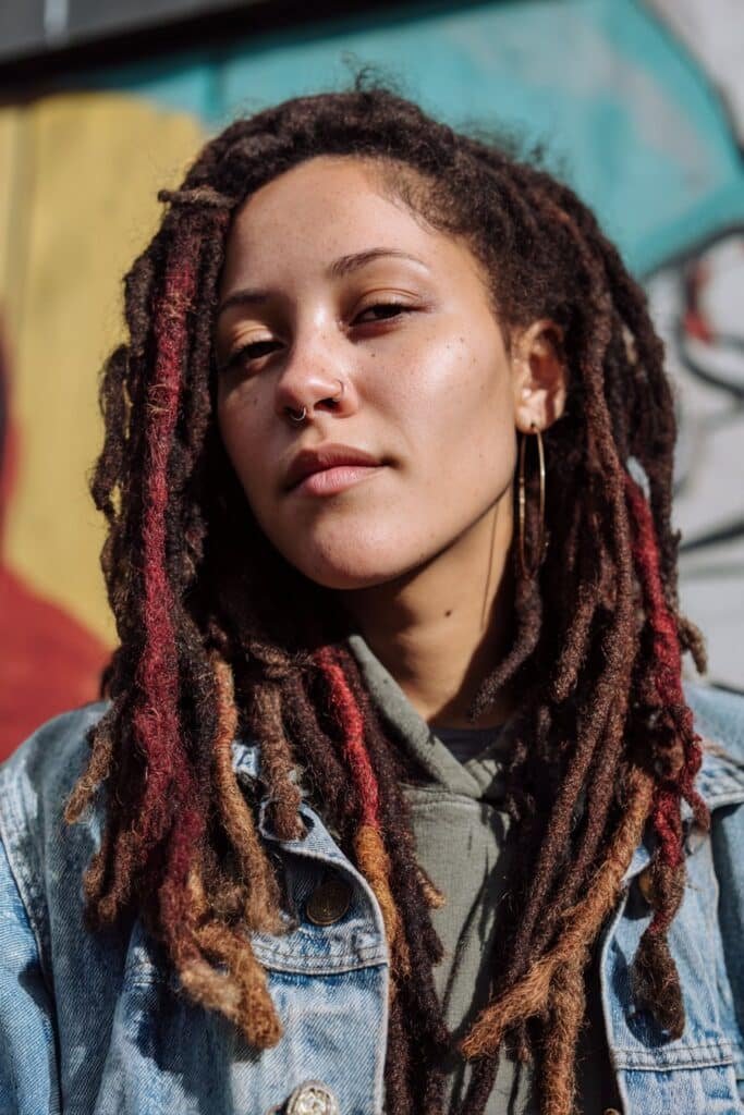 Chest-up front-facing image of a woman in her 20s wearing locs accented with red streaks, emphasizing texture and contrast against an urban mural.