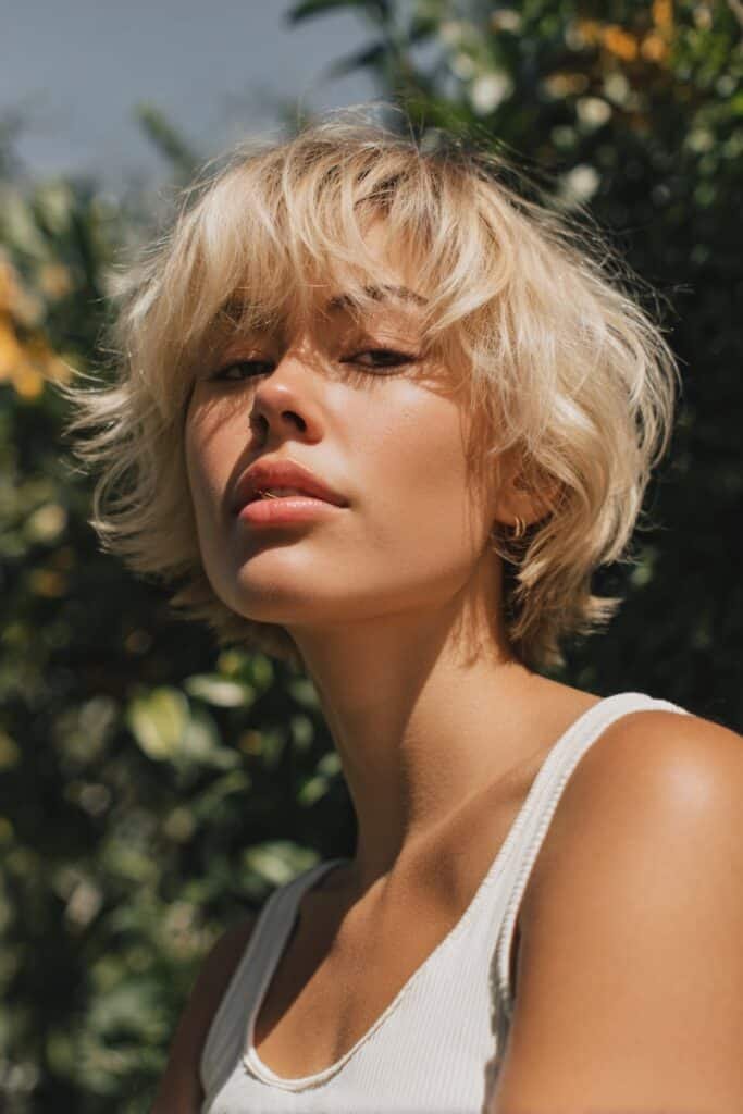 A woman with thin fine hair wearing a shaggy blonde lob, captured outdoors with blurred garden greenery in the background.