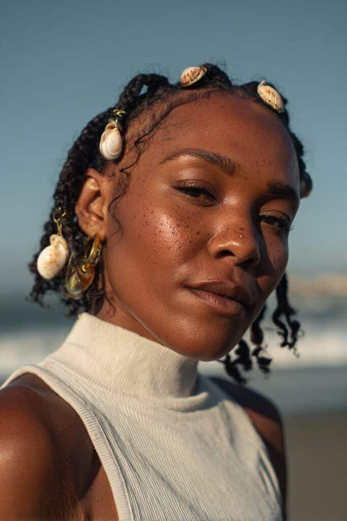 Woman in her 20s wearing a light turtleneck with short Fulani braids decorated with shell accessories at a beachside location