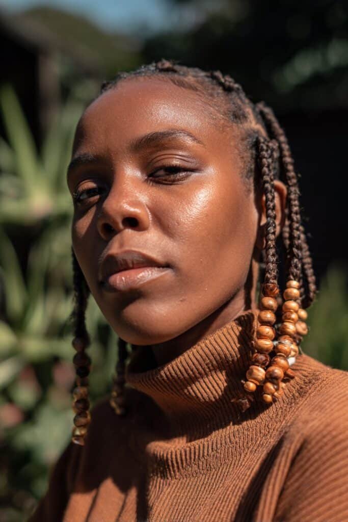 Woman in her 20s wearing an earth-tone turtleneck with short Fulani braids decorated with wooden beads in an outdoor garden setting.