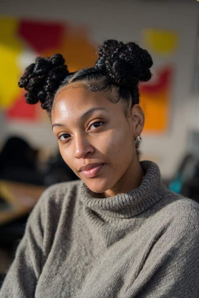 Playful woman wearing a cozy turtleneck with short Fulani braids styled into double buns in a colorful creative studio.