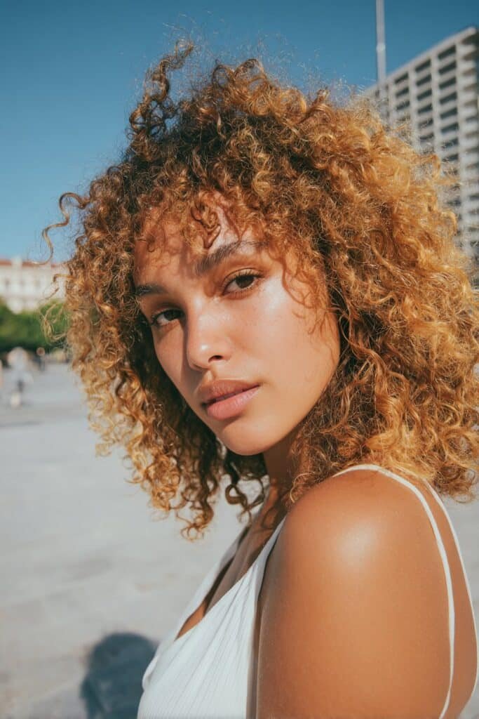 Young woman in her 20s with fine thin curly hair in a shoulder-length cut with soft highlights, white summer outfit, standing in a sunny city plaza.