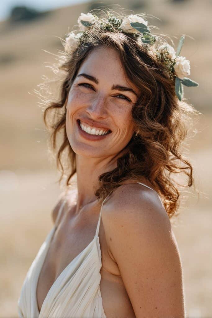 Bride in her 20s wearing soft curled wedding hair topped with a floral hair crown, photographed in an outdoor meadow wedding scene.