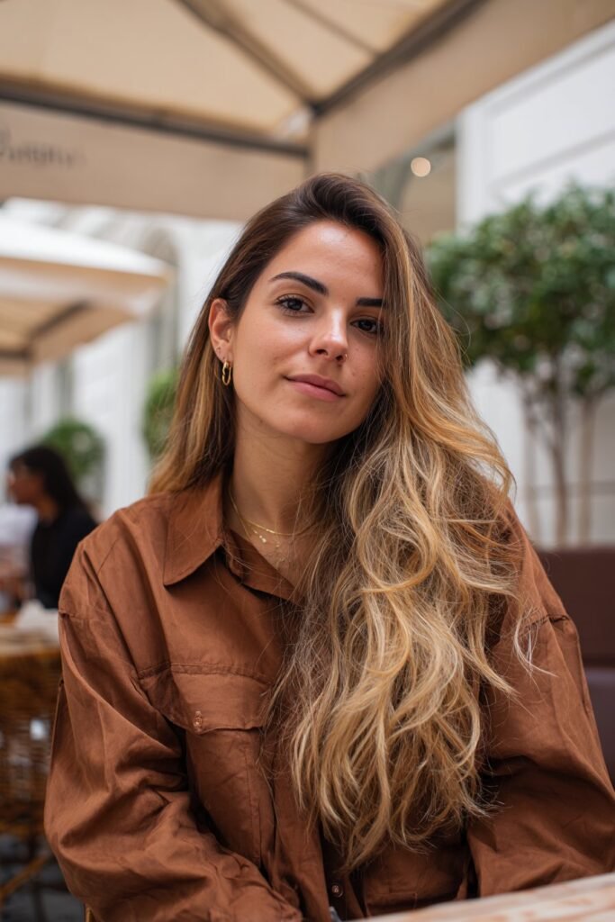 Woman in her 20s with toasted butter blonde hair, deeper warm blonde tones set against an outdoor café patio.