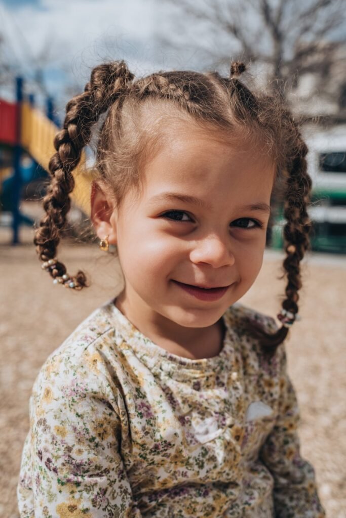 A toddler girl with side-swept lemonade braids adorned with beads, photographed outdoors in a sunny park setting.