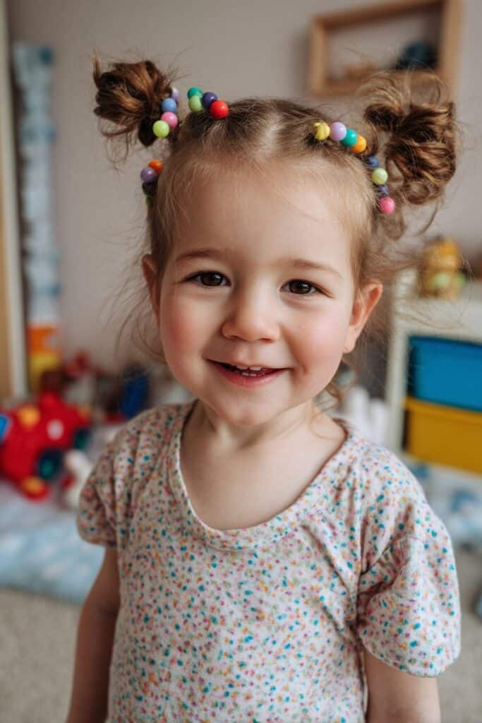 A toddler girl with twin bun braids decorated with colorful beads, photographed in soft nursery lighting with a playful pastel background.