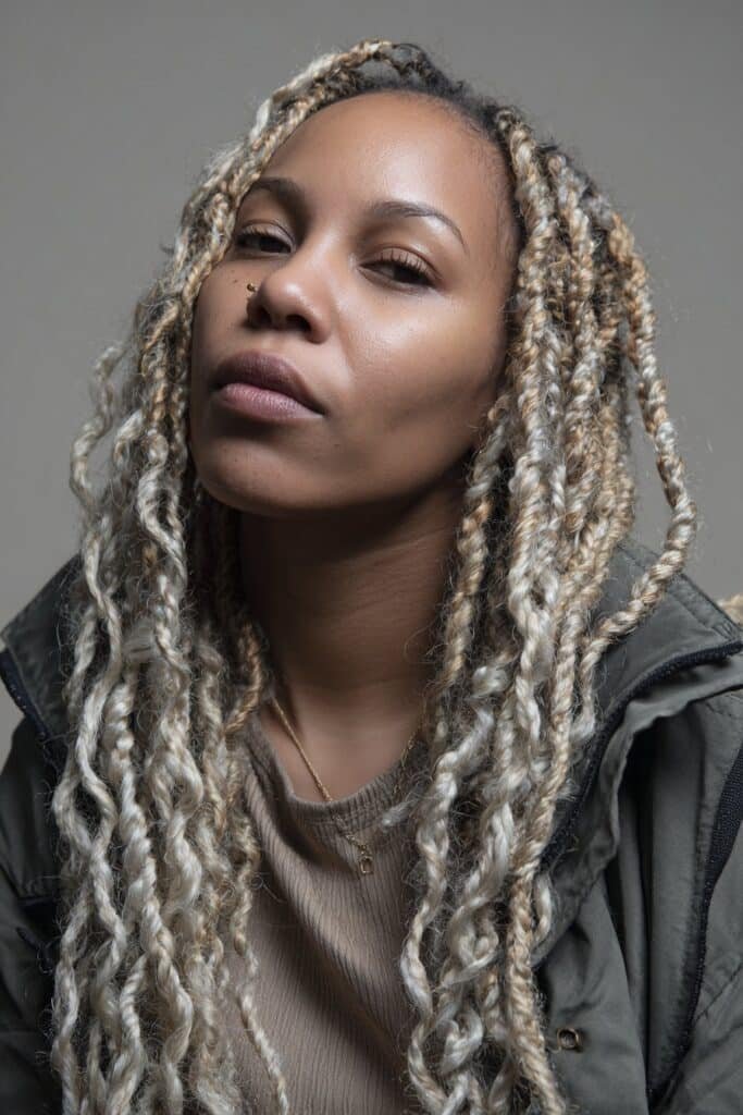 Woman in her 20s featuring twists and braids with platinum tones, intricately styled icy blonde braids in a warm studio backdrop.