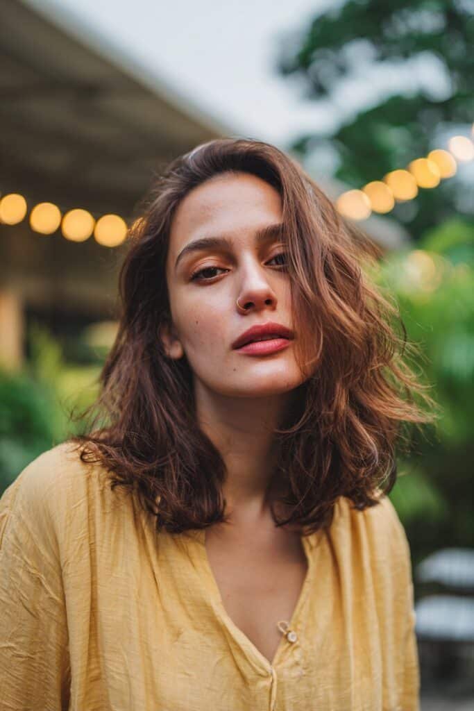 Young woman with thin fine wavy hair in a voluminous medium-length cut with side part on a garden patio with string lights.