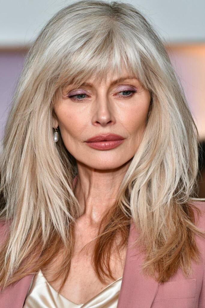 Older woman with a layered shag haircut and wispy bangs in ash blonde hair.