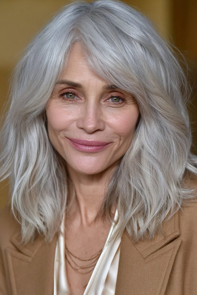 Elegant woman with salt and pepper waves and soft curtain bangs wearing a camel blazer in a beige studio setting.