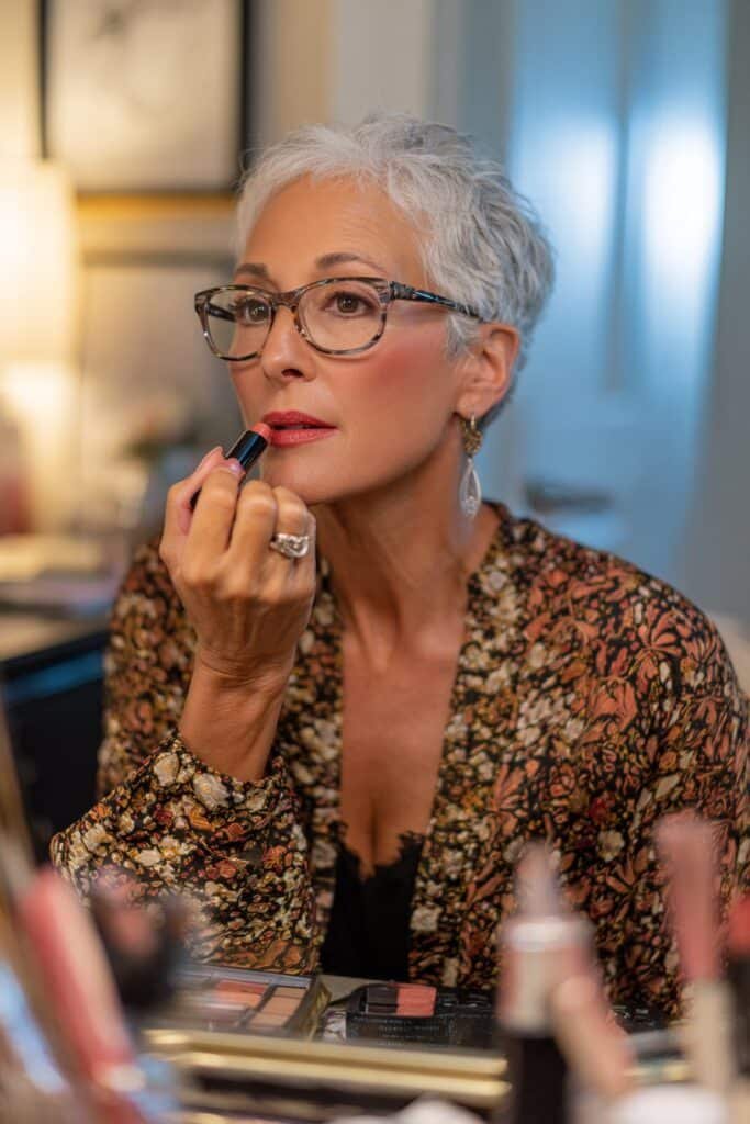 Mature woman with glasses applying a soft lipstick shade that balances her makeup.