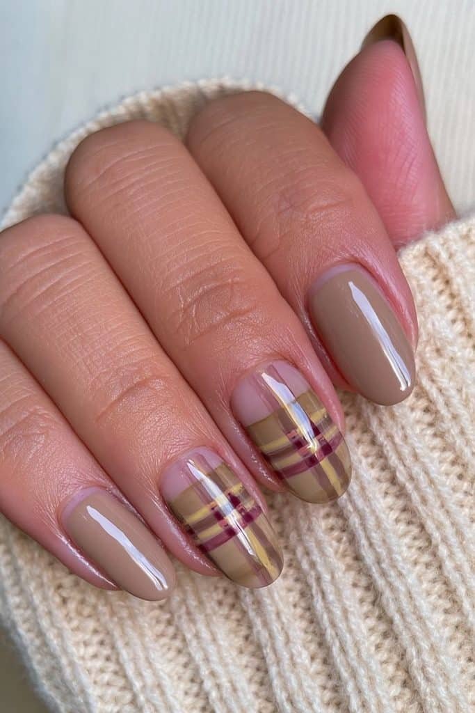 Short oval Thanksgiving nails with warm nude and brown tones and a burgundy plaid accent.