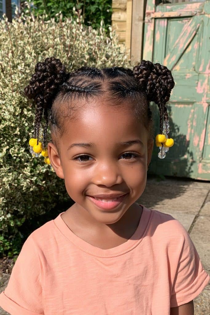 Kids braided buns with two symmetrical buns, neat sections, and bright yellow and clear beads.