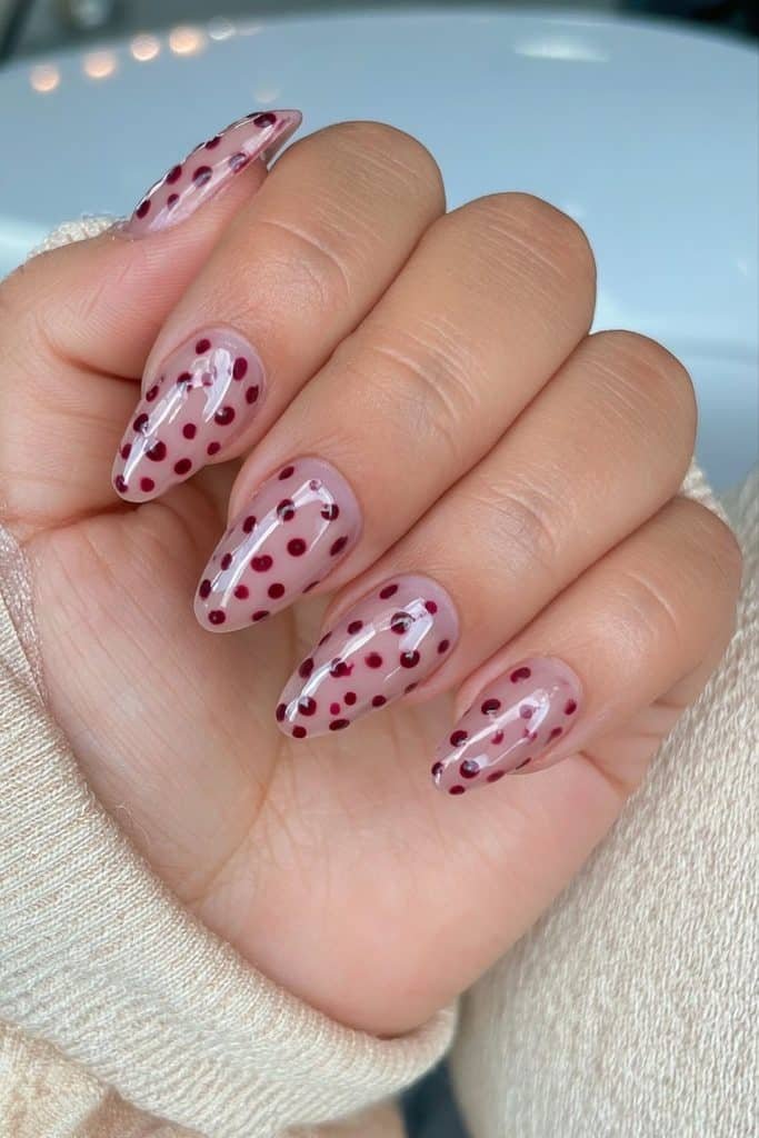 Sheer nude nails with delicate burgundy polka dots in varied sizes and glossy shine