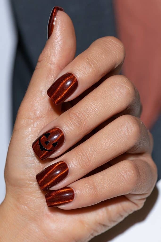 Orange cat eye Halloween nails with a single accent nail featuring black jack-o’-lantern eyes and a grin.