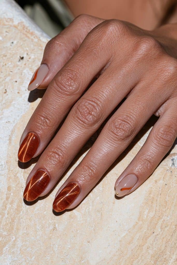 Orange cat eye nails paired with two nude accent nails featuring tiny gold or orange details.